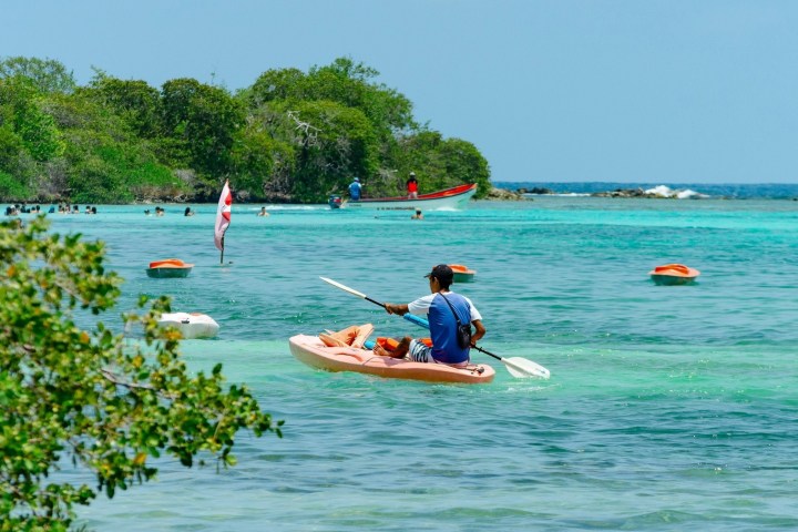 Person kayaking in clear turquoise water near a beach with trees in the background.