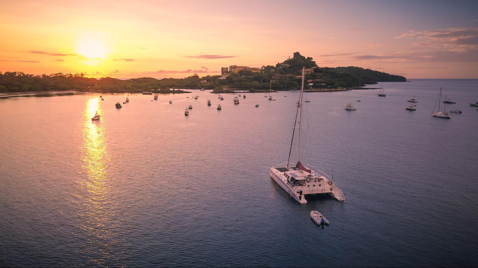 Sunset over calm bay with numerous boats anchored, including a large catamaran in the foreground.