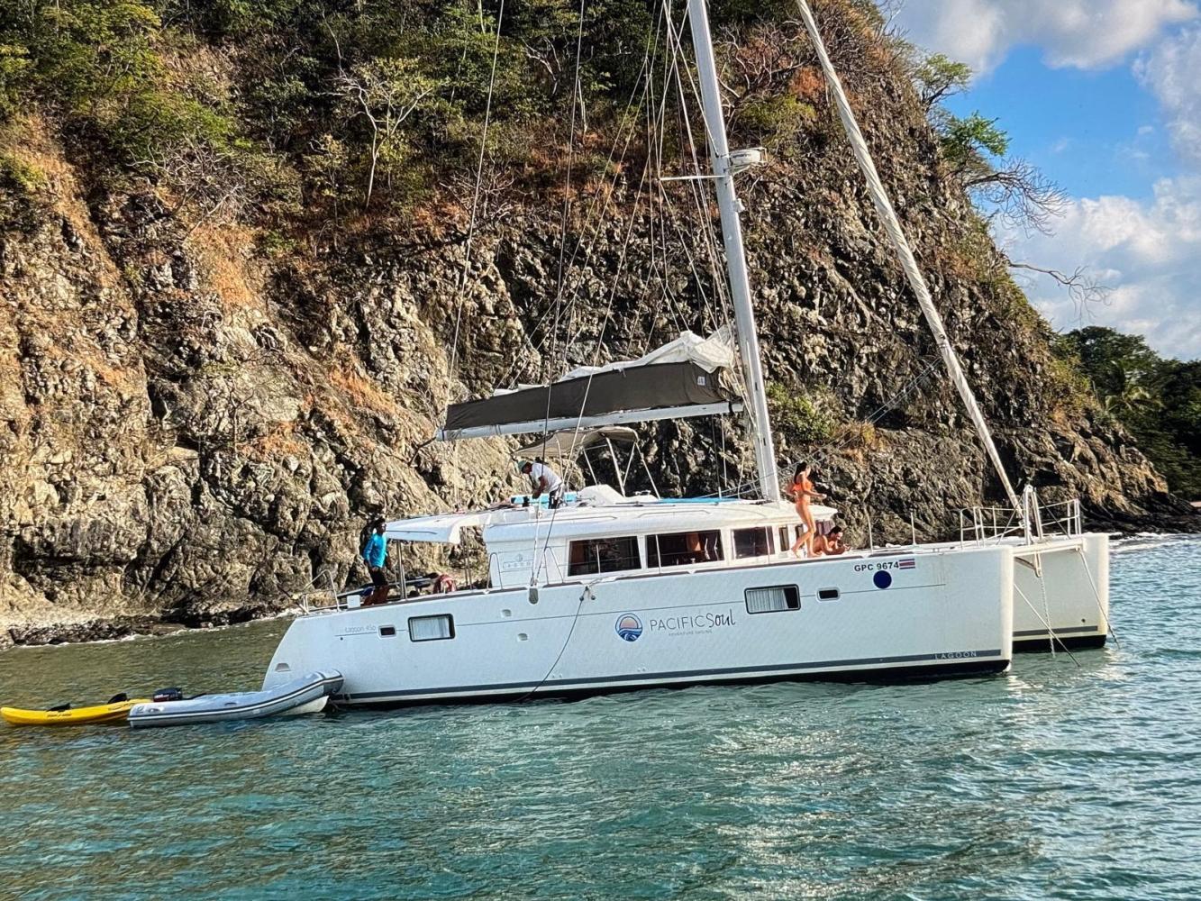 Catamaran named 'Pacific Soul' with people on board near rocky shore.